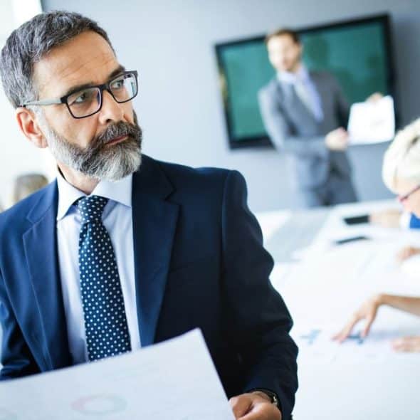 A businessman with glasses and a beard stands in a meeting, holding a document.