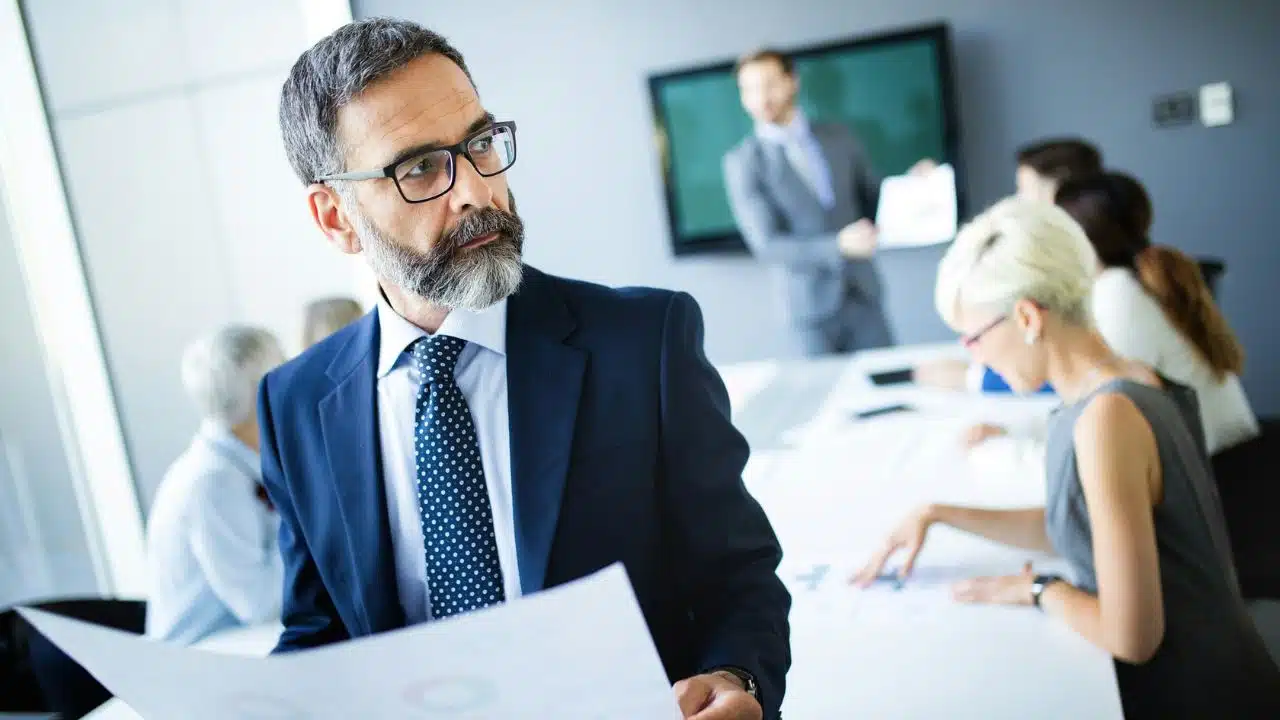 A businessman with glasses and a beard stands in a meeting, holding a document.
