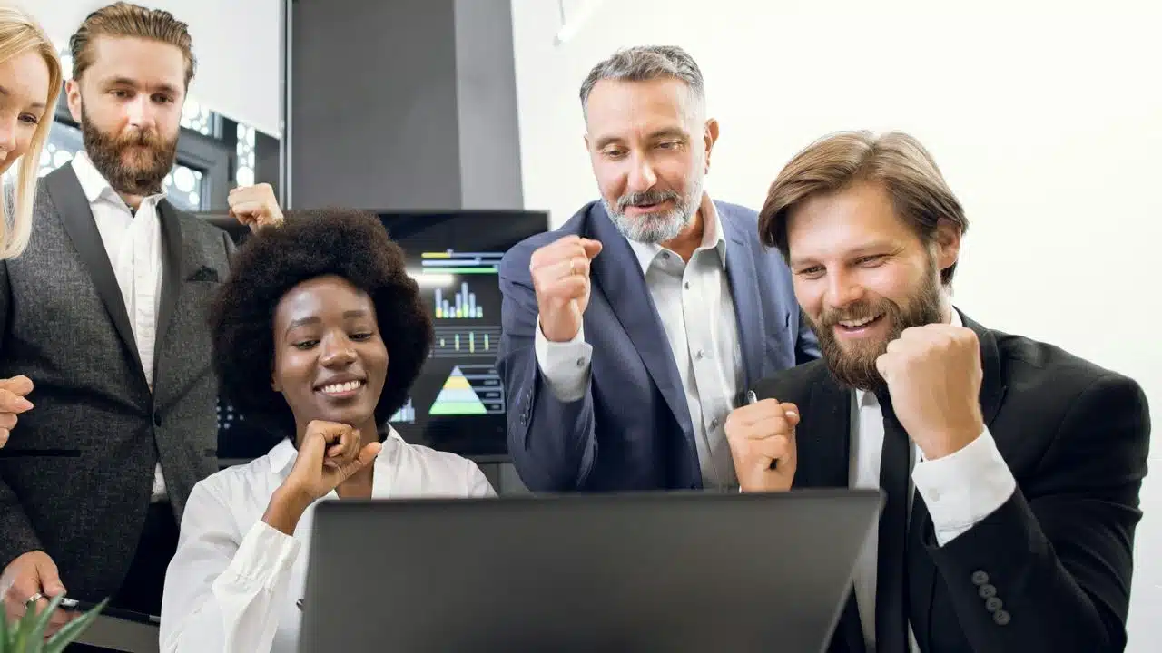 A diverse group of five coworkers celebrates a success while looking at a laptop.