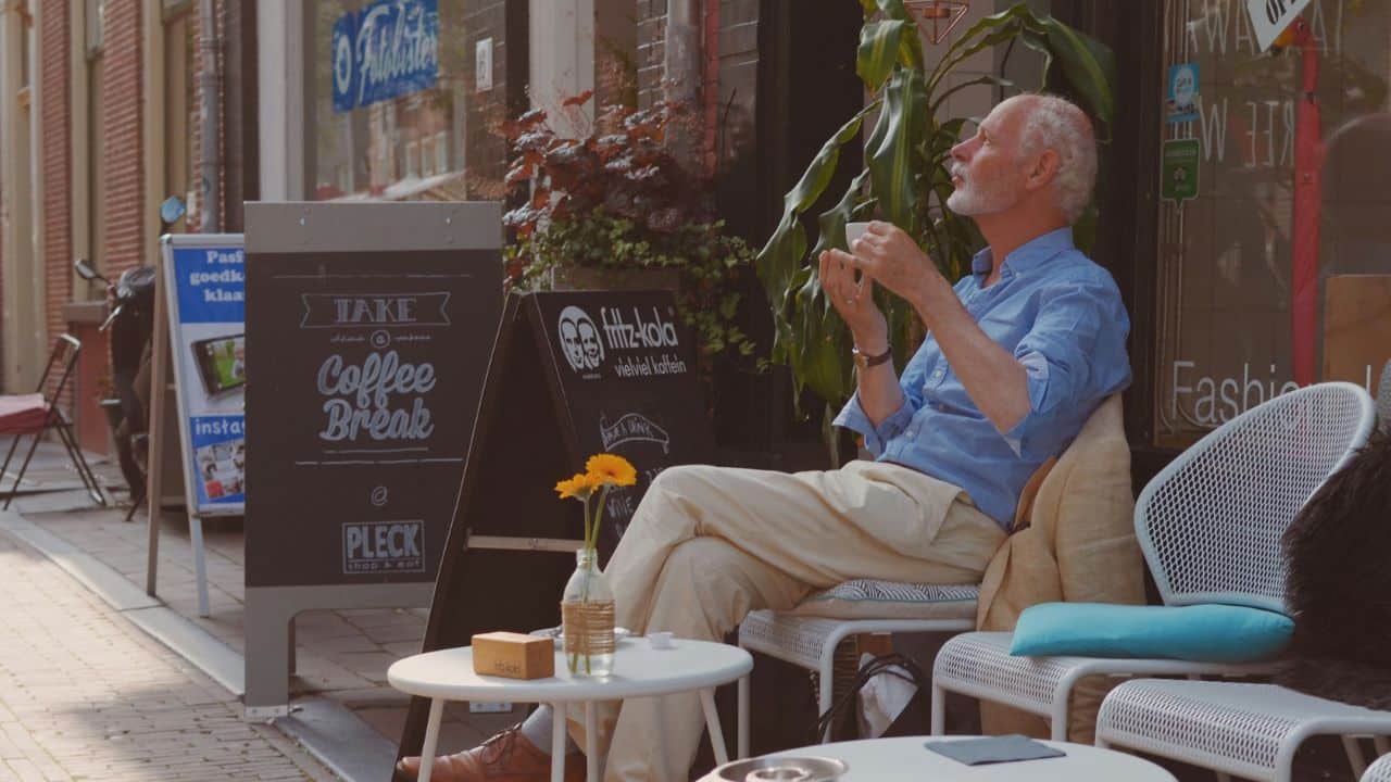 A mature man in a blue shirt relaxes with a coffee outside a cafe.