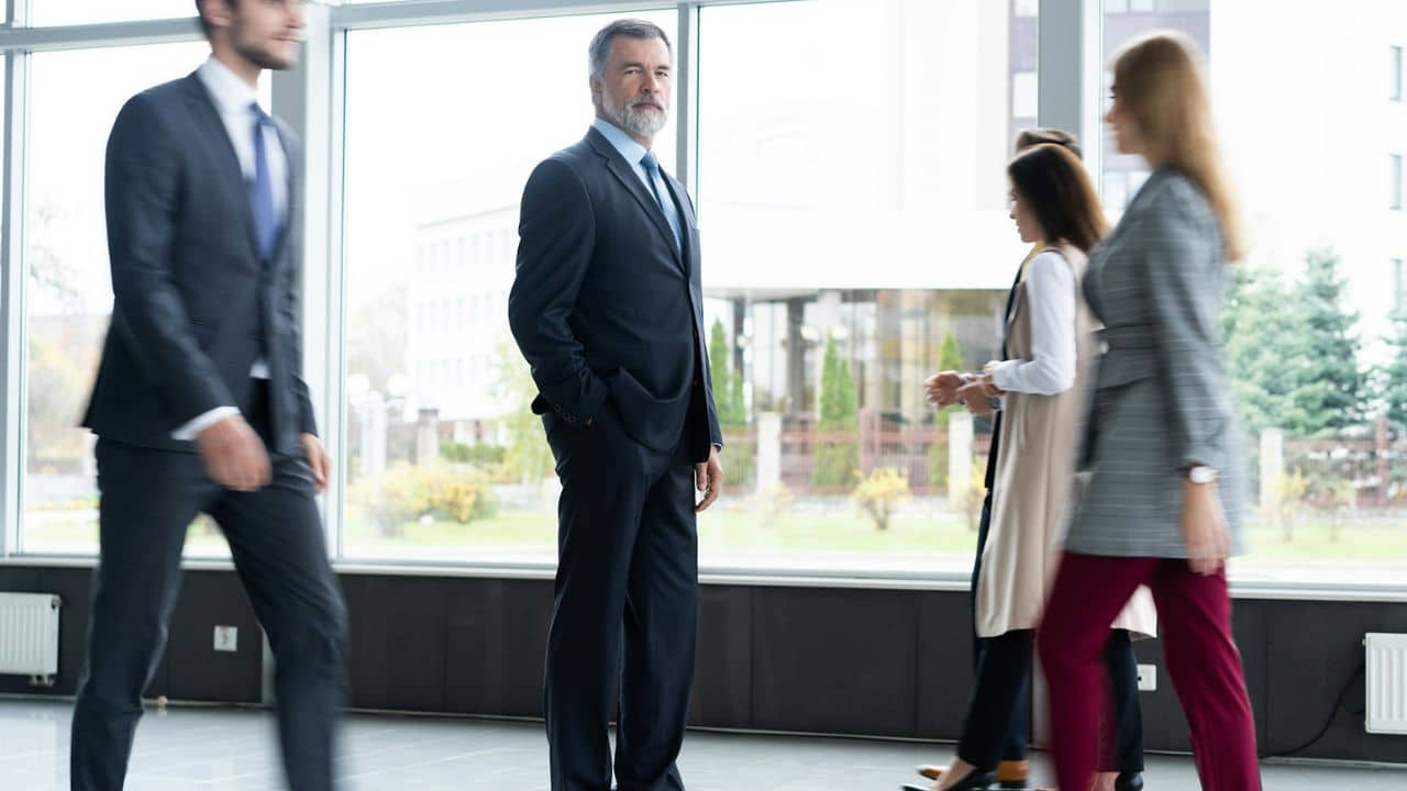 A confident businessman stands still in an office hallway while people walk by.