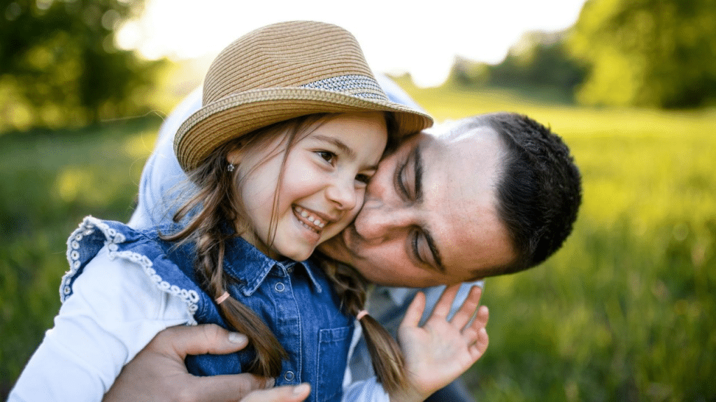 A smiling girl in a denim jumper and straw hat is hugged by a man in a grassy field.