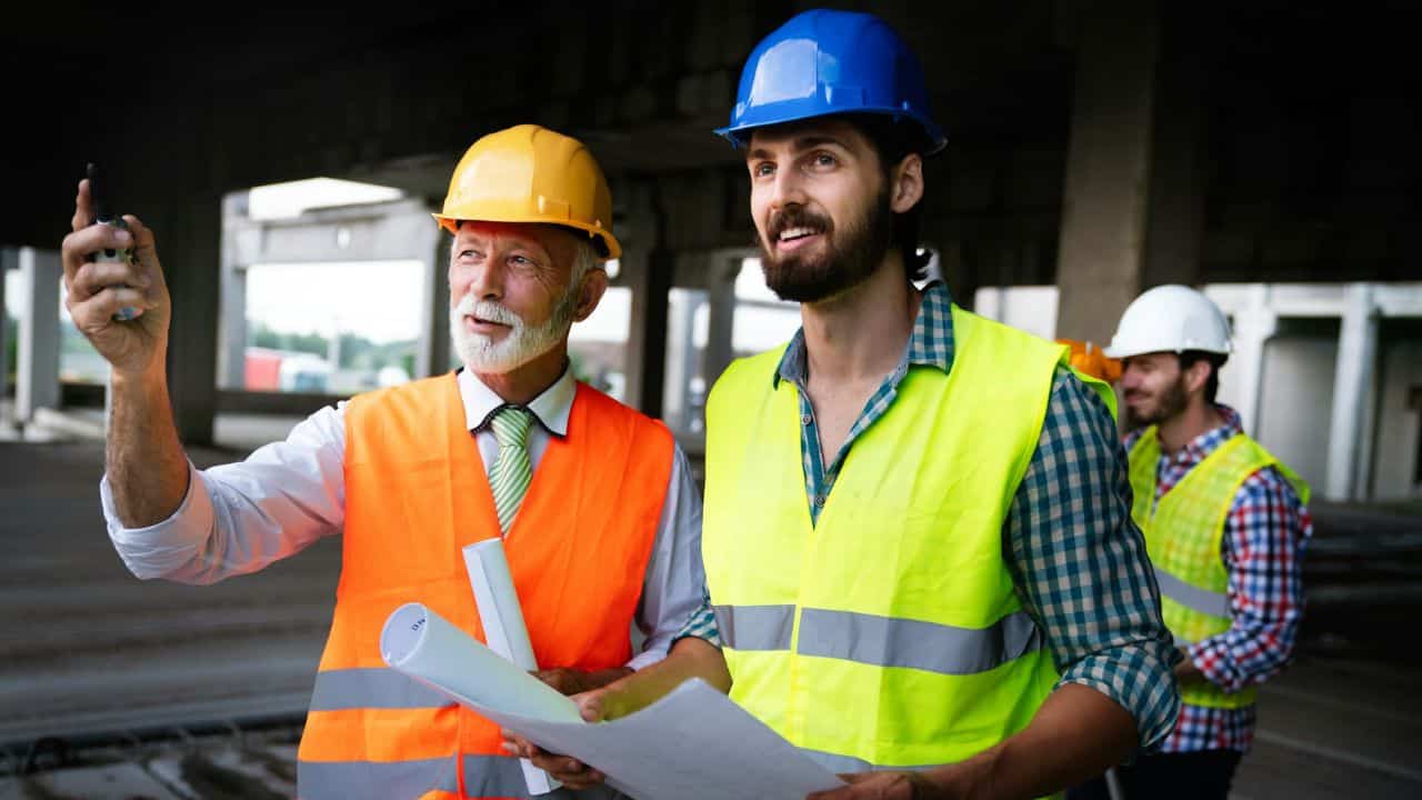 Two male construction workers in hard hats look at a document together.