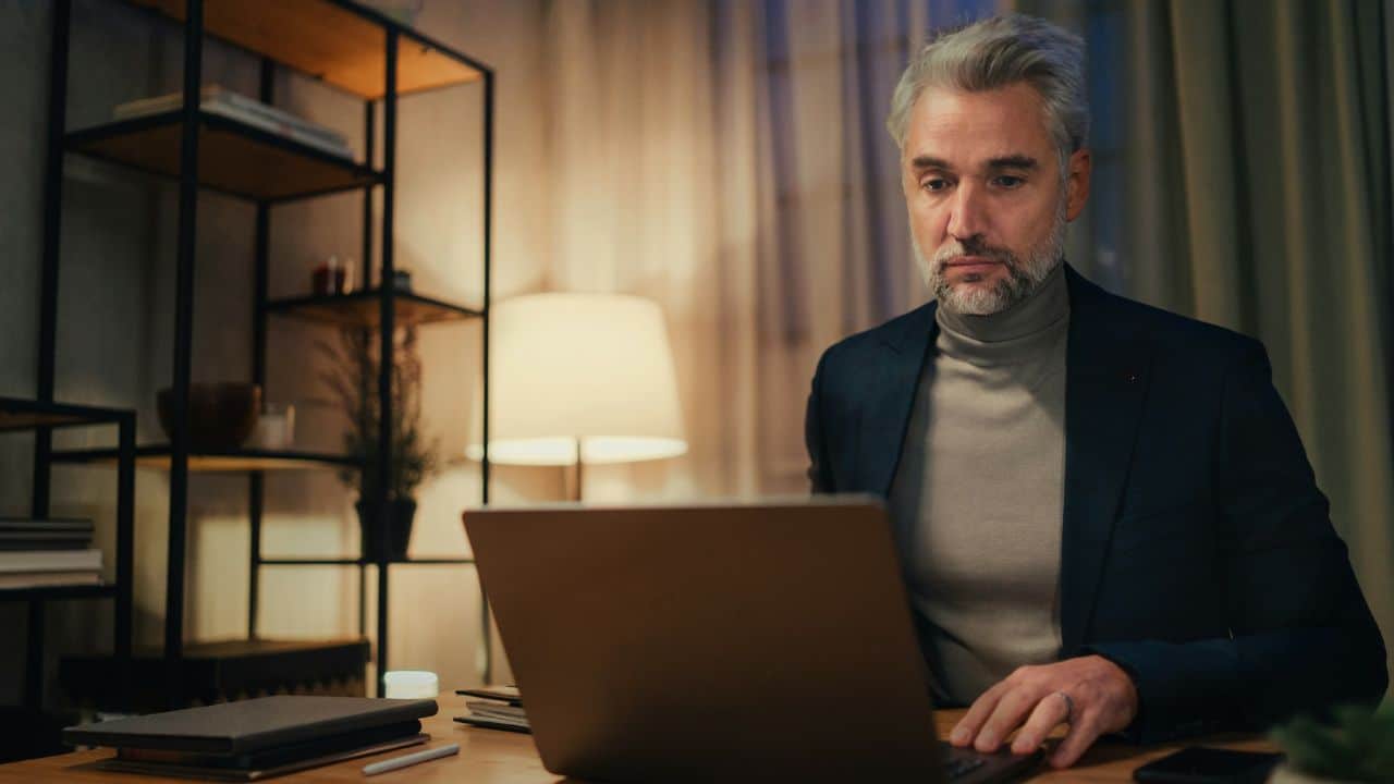 A handsome mature man with a beard works intently on his laptop at home.