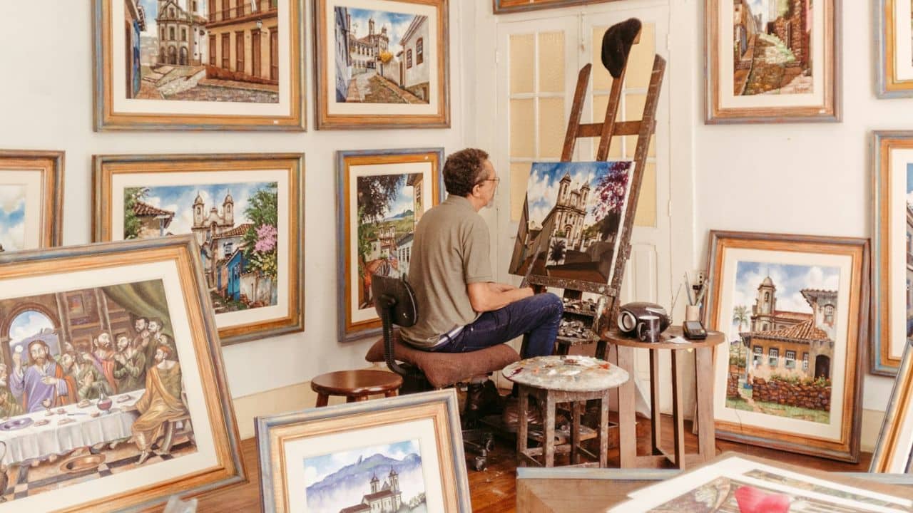 A painter sits on a stool and works on a canvas surrounded by many finished paintings.