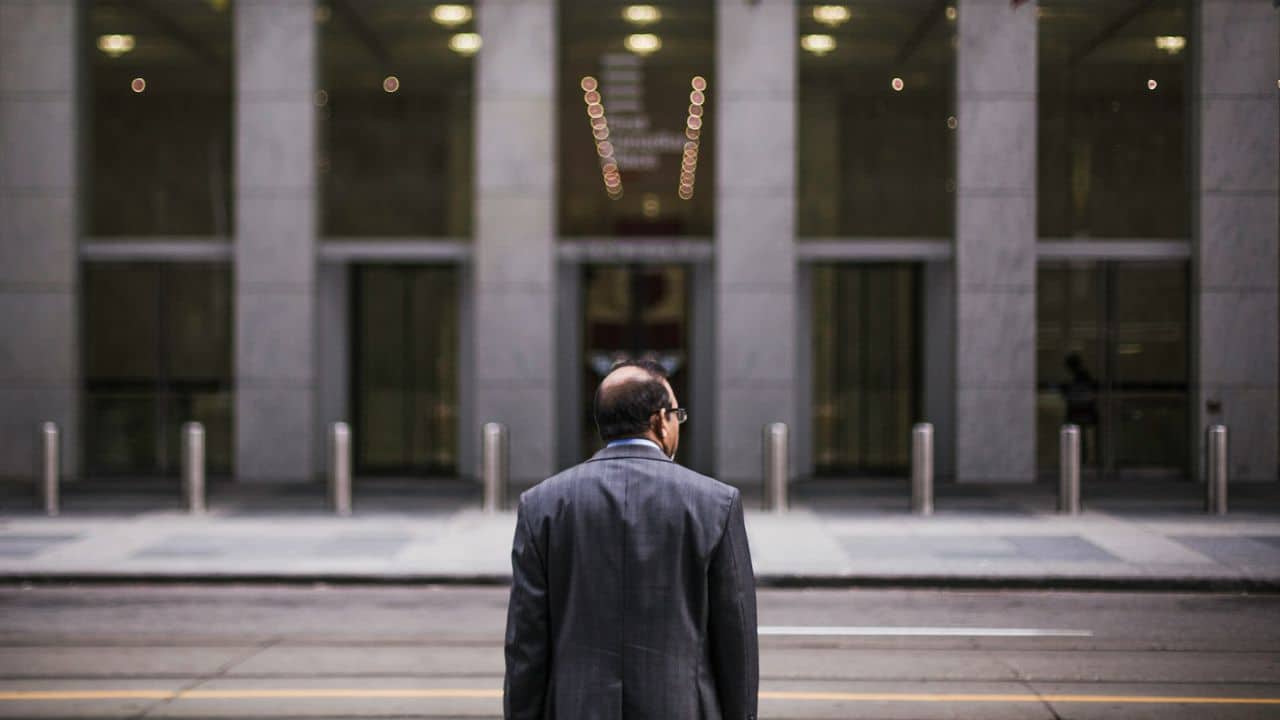 A man in a suit stands in the street, looking at a modern building.