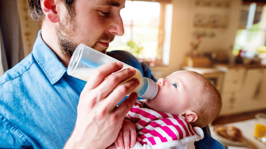 A man with a beard holds and feeds a baby from a bottle.