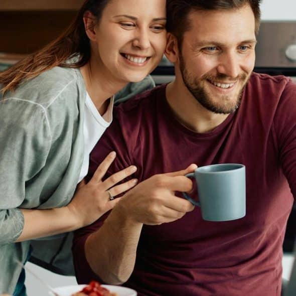 A smiling couple looks at a laptop together, with the man holding a mug.