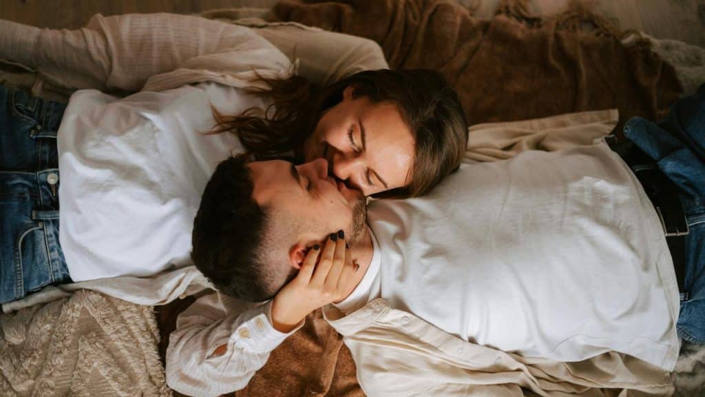 A couple lies on a blanket, smiling and sharing a kiss.