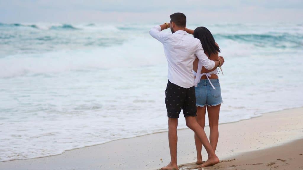 A couple walks barefoot on the beach, embracing as they look at the ocean.