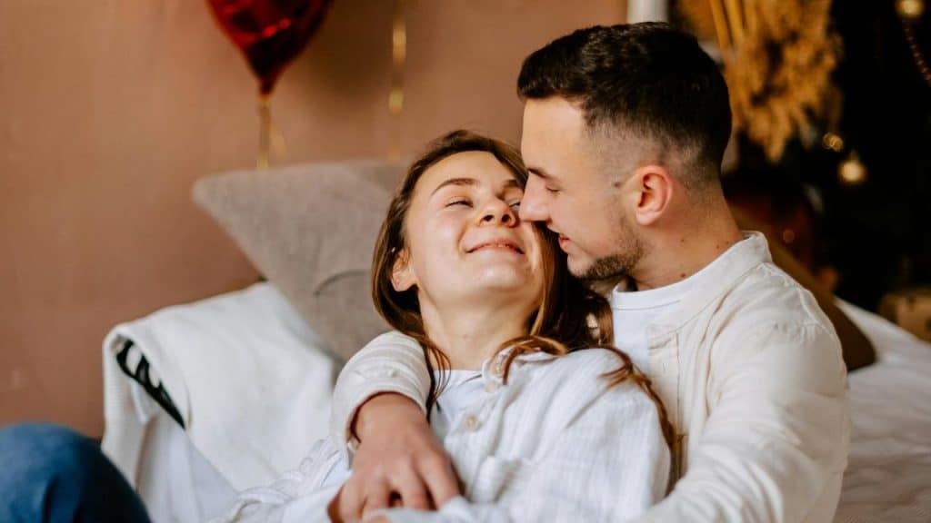 A smiling couple cuddles together indoors, looking at each other affectionately.