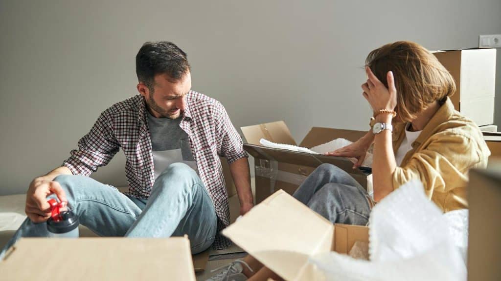 A couple sits on the floor surrounded by moving boxes, unpacking belongings.
