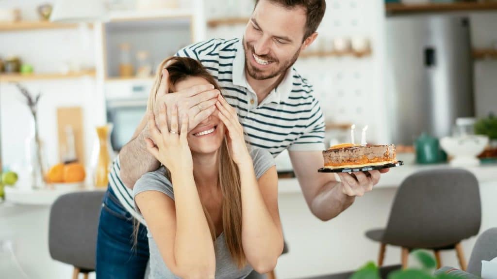 A man surprises a smiling woman with a birthday cake while covering her eyes.