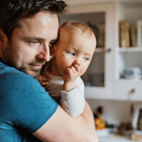 A man with a beard is holding and looking at a baby in his arms.