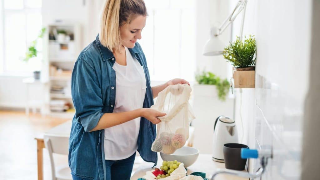 A woman unpacks fresh produce from a reusable bag in the kitchen.