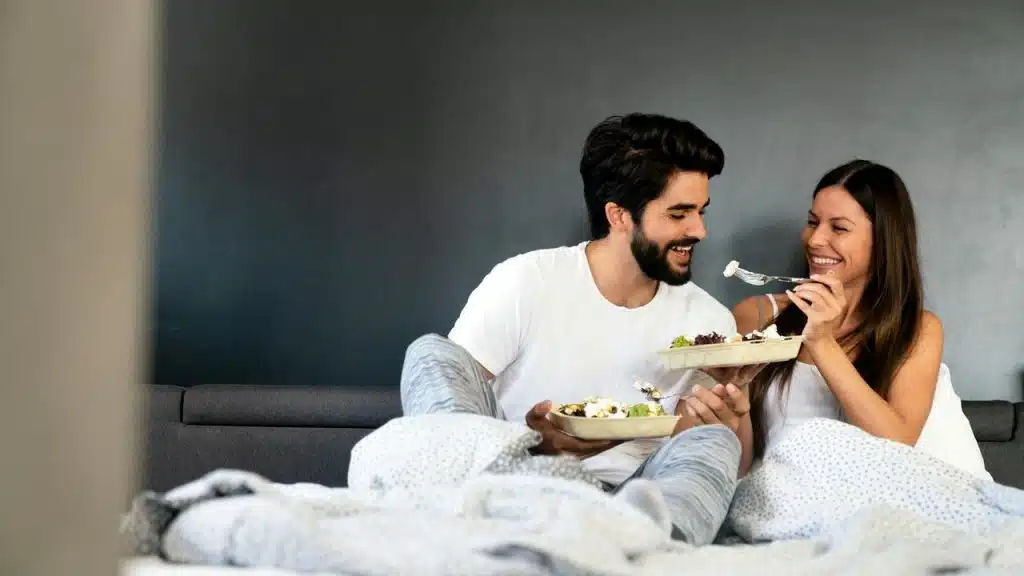 A smiling couple enjoys breakfast in bed, feeding each other.
