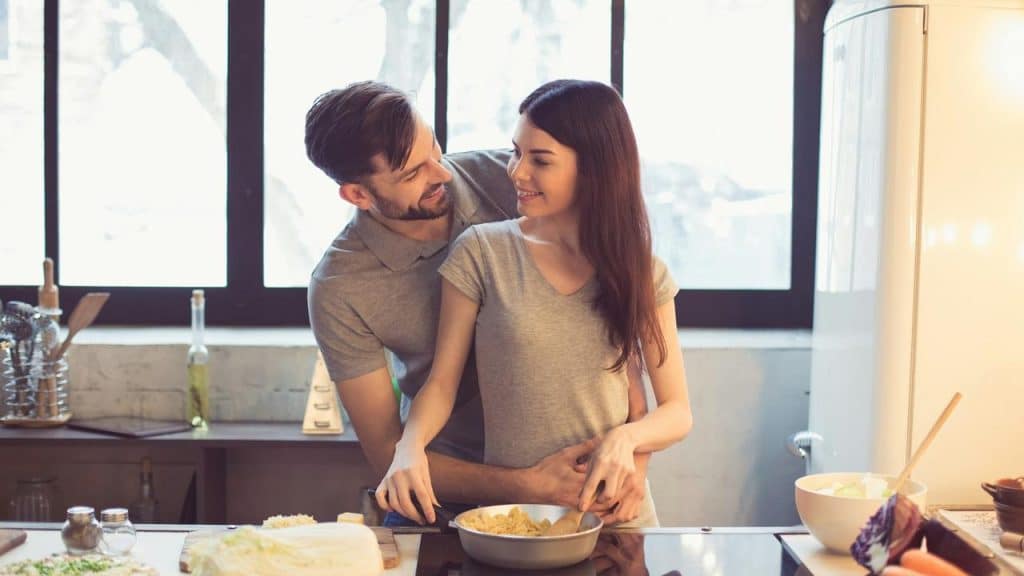 A couple smiles while cooking together in the kitchen.