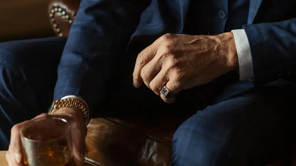 A man in a navy suit wearing a gold watch and ring while holding a glass of whiskey.