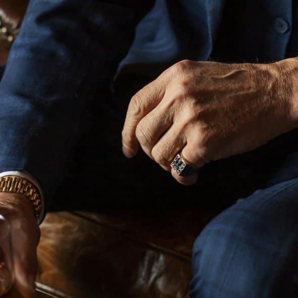 A man in a navy suit wearing a gold watch and ring while holding a glass of whiskey.