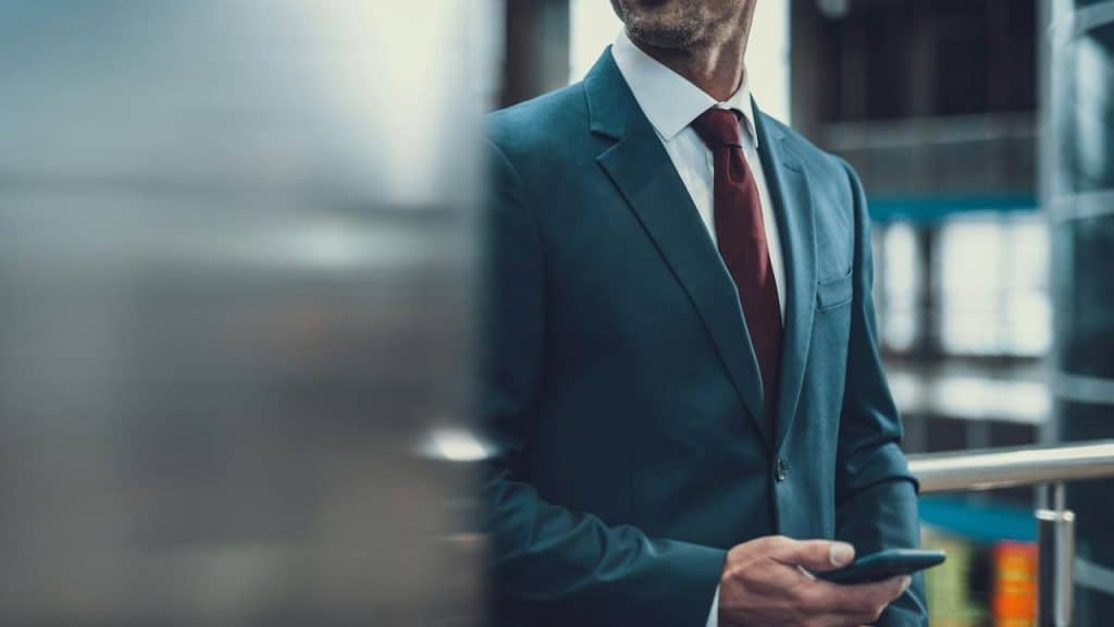 A man in a dark suit with a red tie holding a smartphone indoors.
