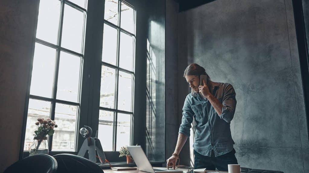 A man in a denim shirt talking on the phone while working on a laptop.