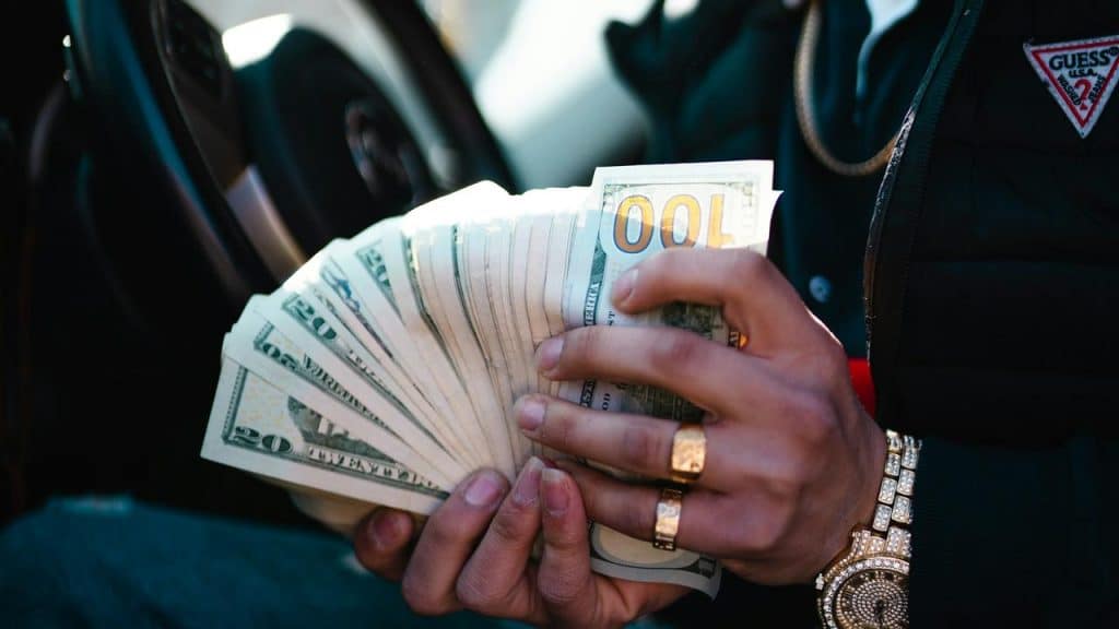A man in jewelry holding a fan of US dollar bills inside a car.
