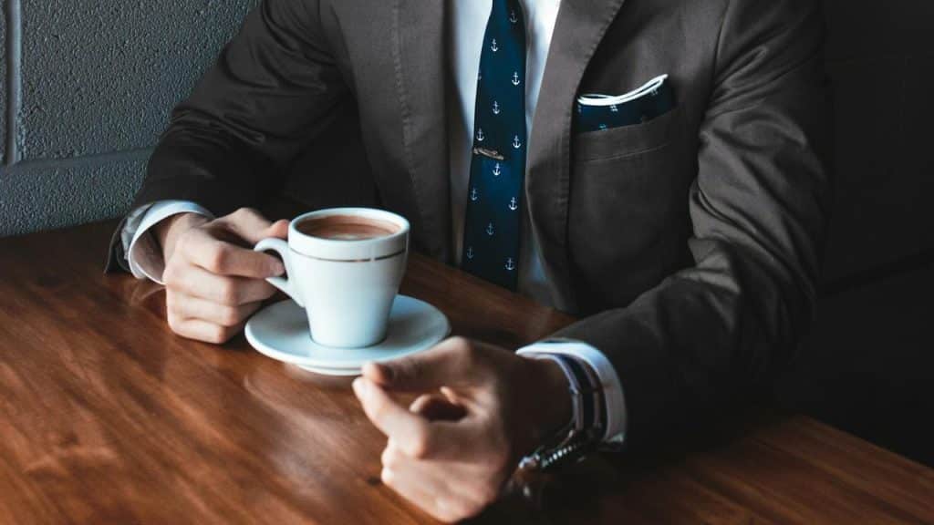 A man in a suit holding a cup of coffee at a wooden table.