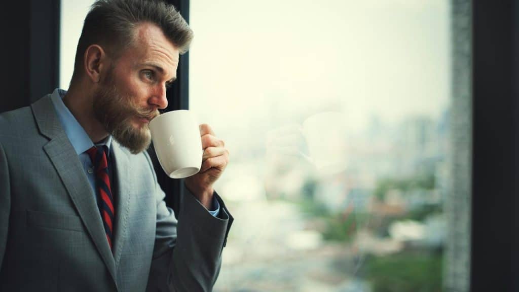 A man in a gray suit drinking from a white mug by a window.