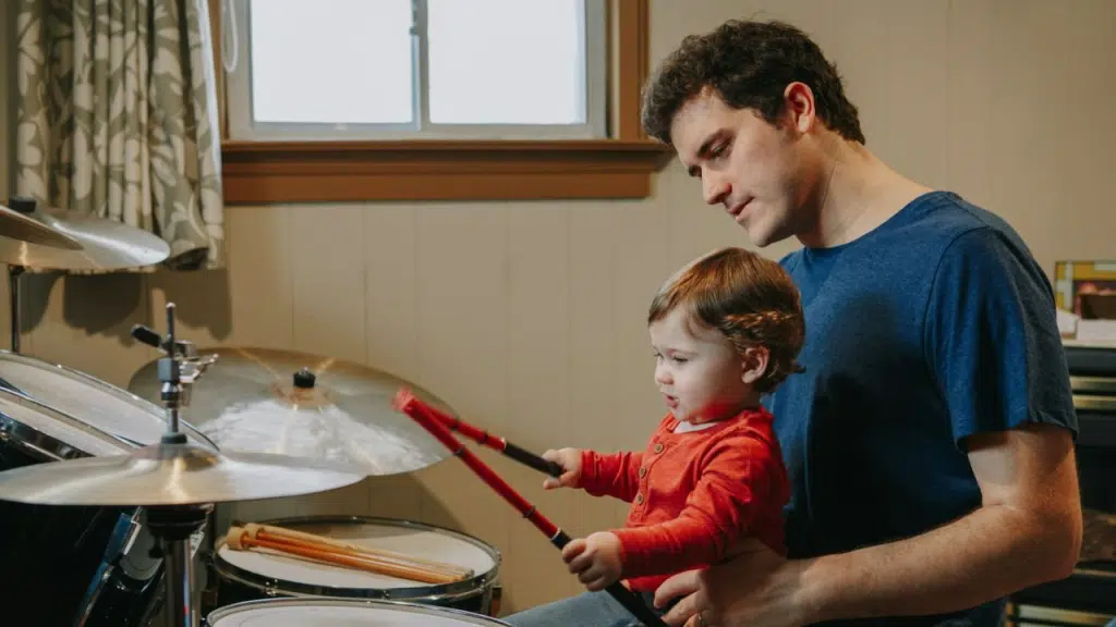 A man is holding a young child in his lap while the child holds two drumsticks over a drum set.