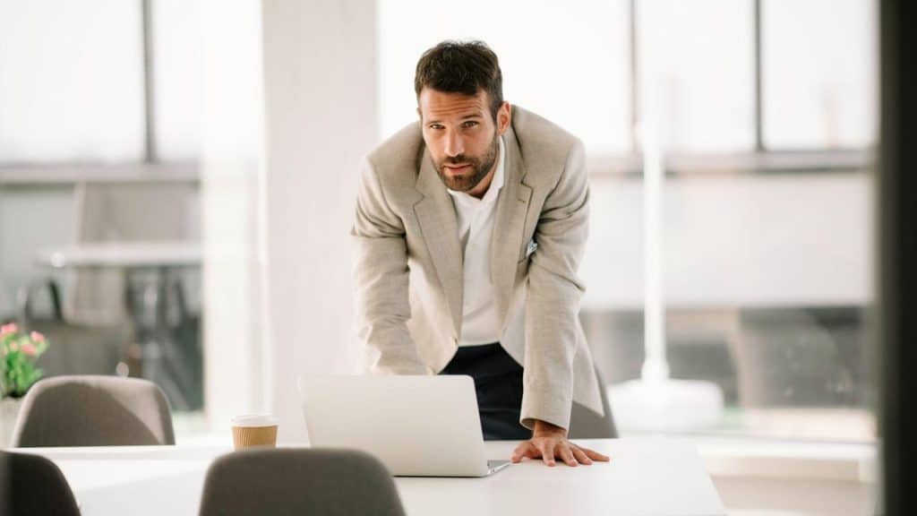 A man in a beige blazer leaning on a table with a laptop in front of him.