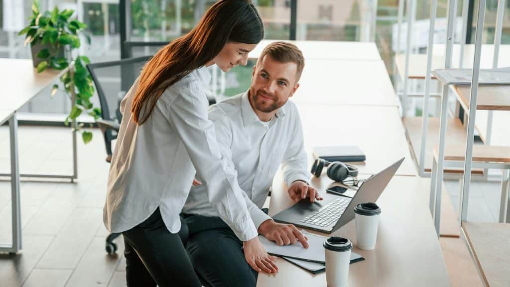 A man working on a laptop while a woman points at a document in an office.