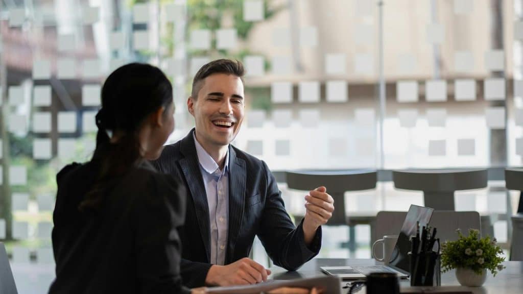 A man smiling and talking with a woman during a meeting in an office.