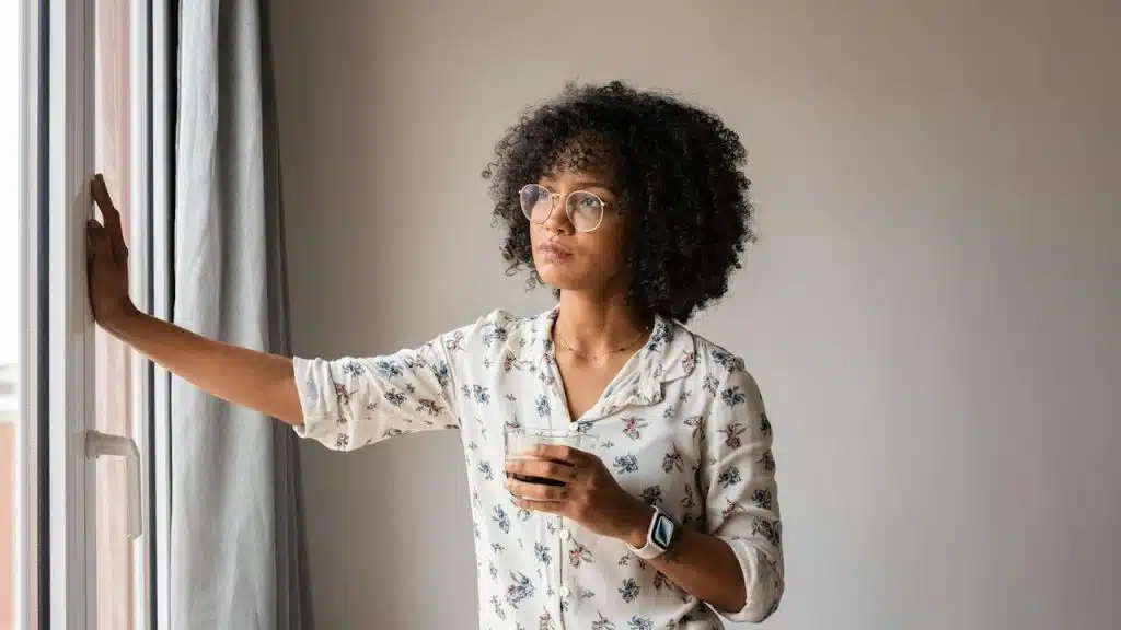 A woman with curly hair and glasses holding a drink while looking out a window.