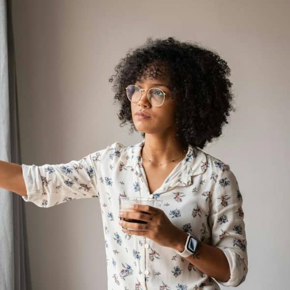 A woman with curly hair and glasses holding a drink while looking out a window.