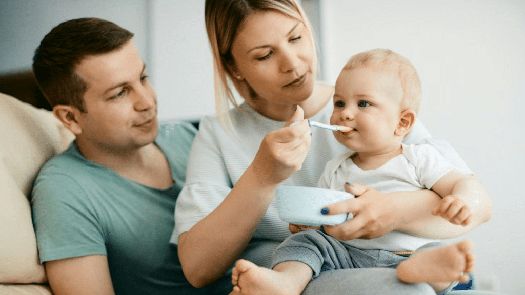 A couple is feeding a baby from a bowl with a spoon while sitting on a couch.