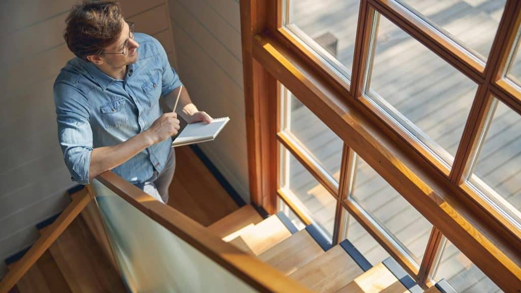 A man standing on stairs by a large window holding a notebook and pencil.
