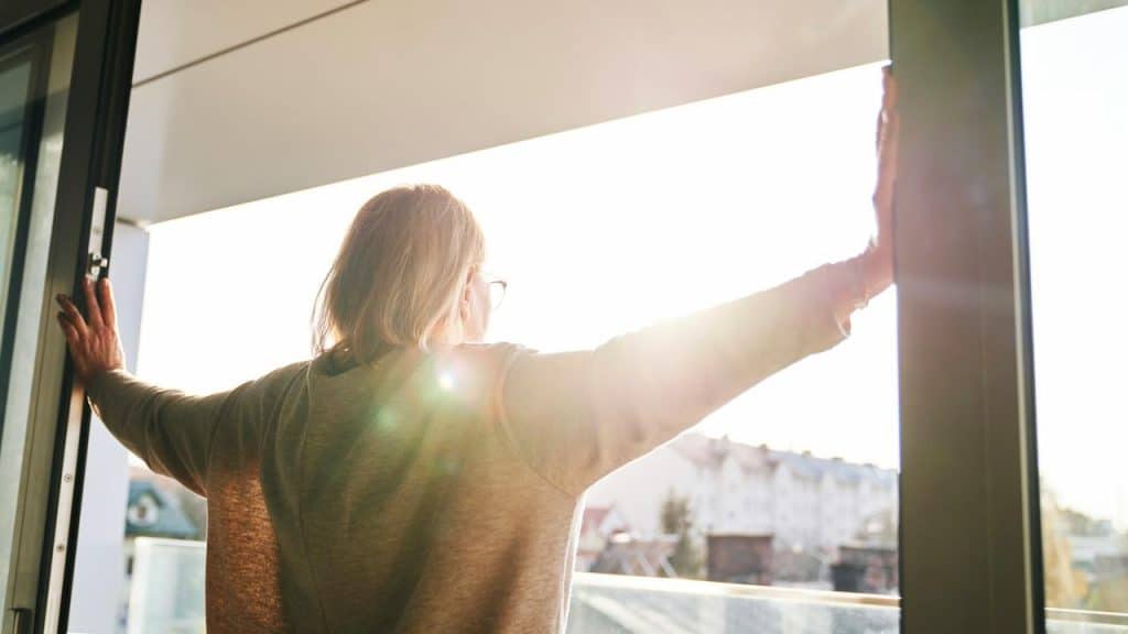A woman standing on a balcony with arms outstretched toward the sunlight.