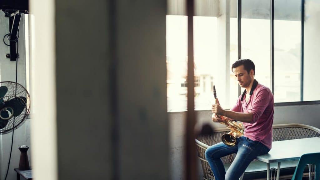 A man sitting indoors by a window playing a saxophone.
