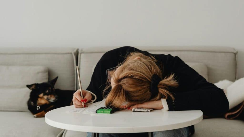 A woman with her head on a table holding a pen, with a dog on the couch.