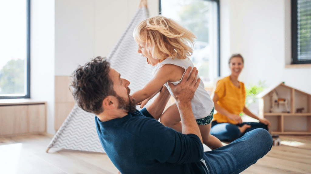 A man is playing with a young blonde child in a room with a teepee. A woman is in the background, sitting on the floor.