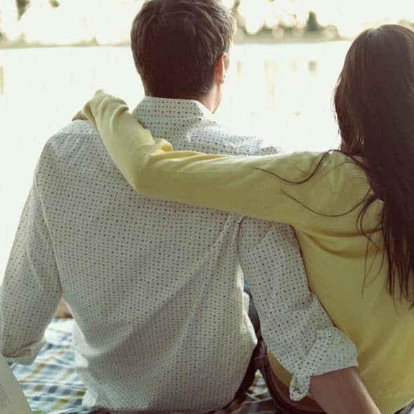 A couple sitting on a blanket by the water, with the woman’s arm around the man.