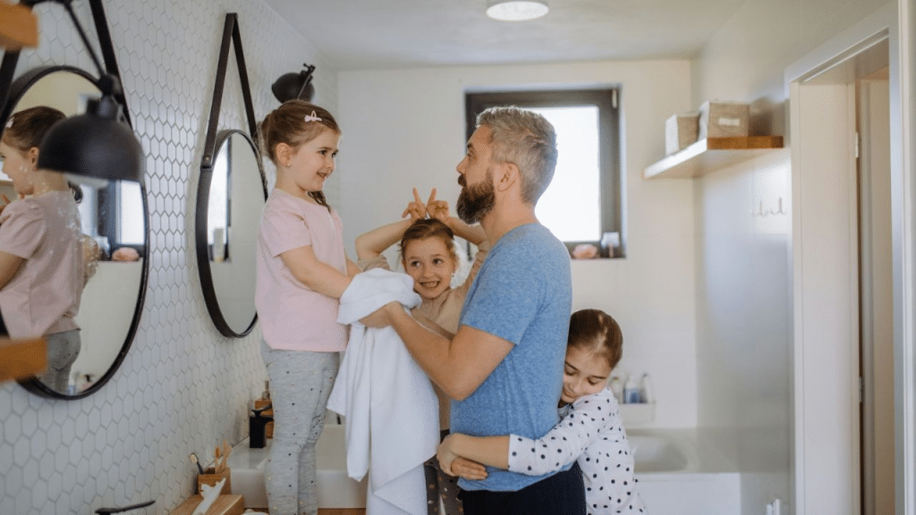 A man is in a bathroom with three young girls who are close to him, with one hugging his leg and another standing on the counter.