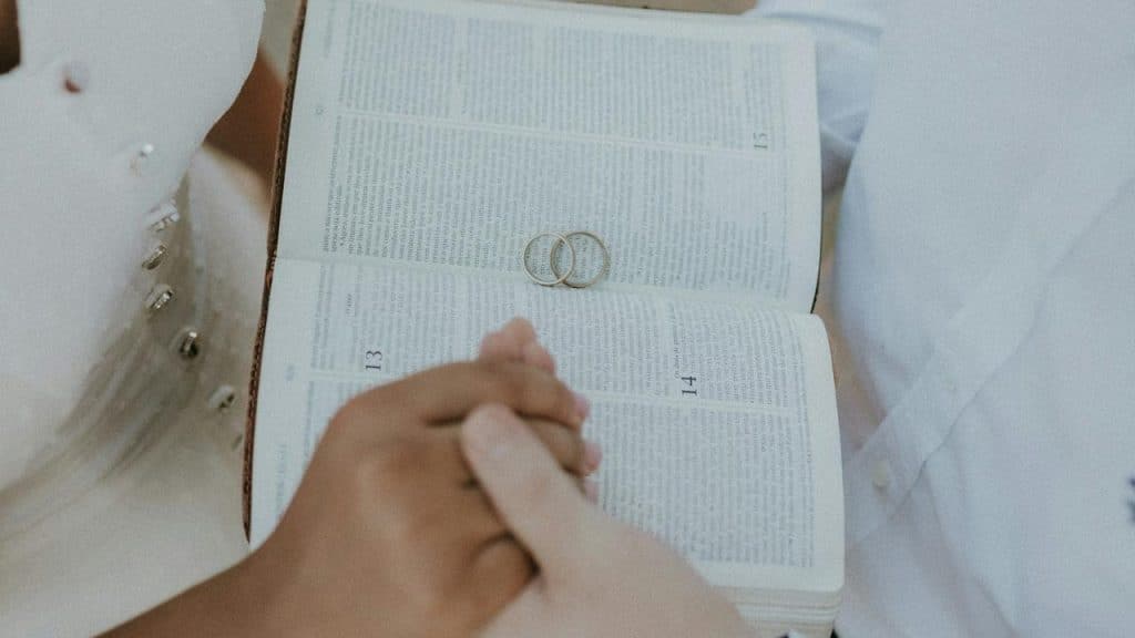 A couple holding hands over an open Bible with two wedding rings placed on the pages.