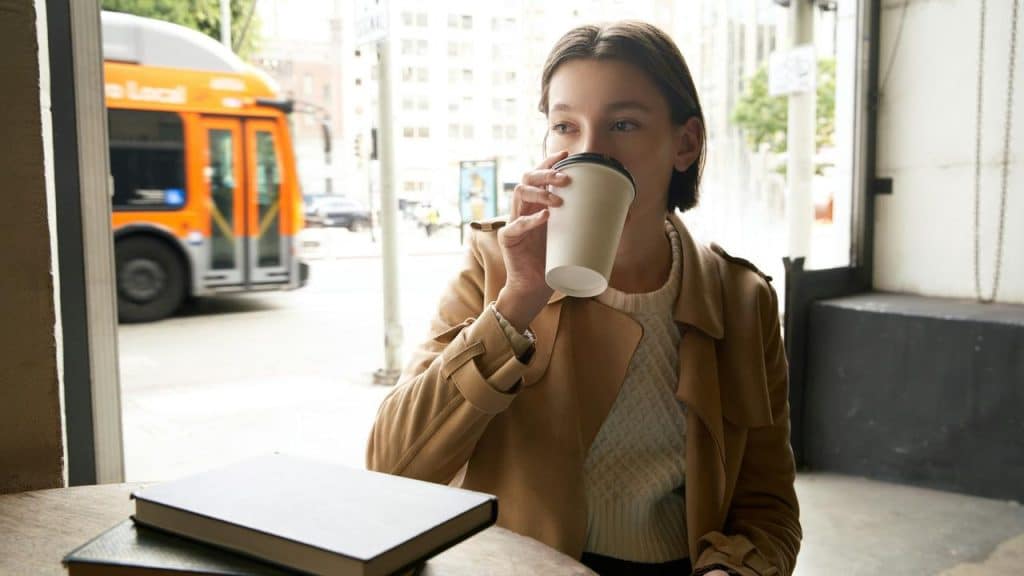 A woman sitting in a café drinking from a paper cup with a notebook on the table.