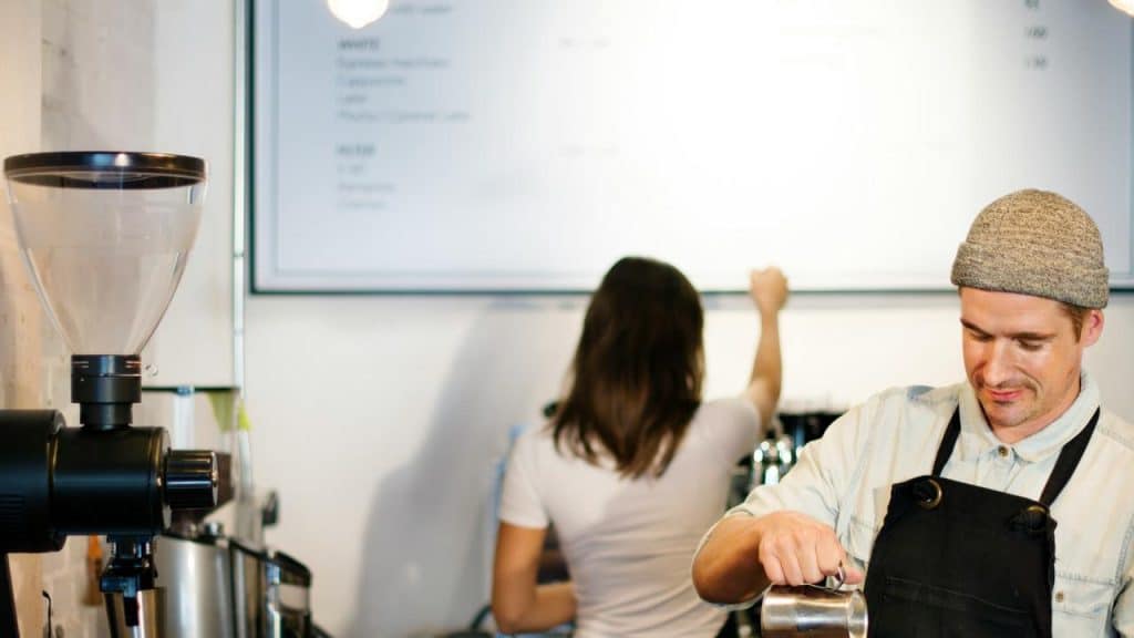 A barista pouring milk with someone updating a café menu board in the background.