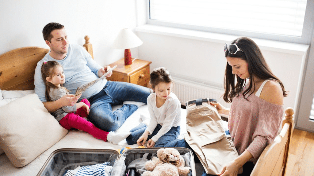 A family of four packing a suitcase in a bedroom with a bed and a window.