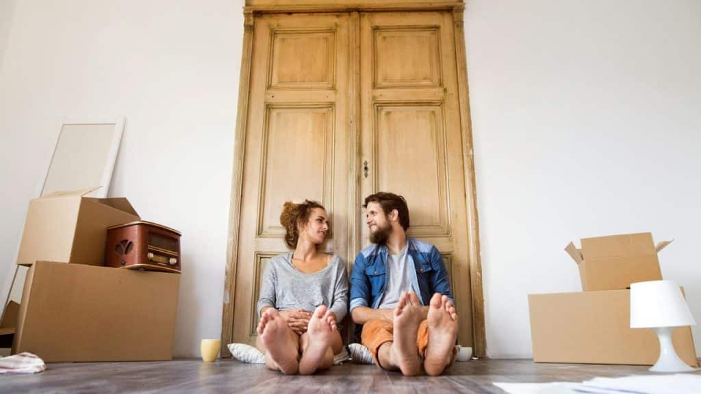 A couple sitting on the floor surrounded by moving boxes, smiling at each other.