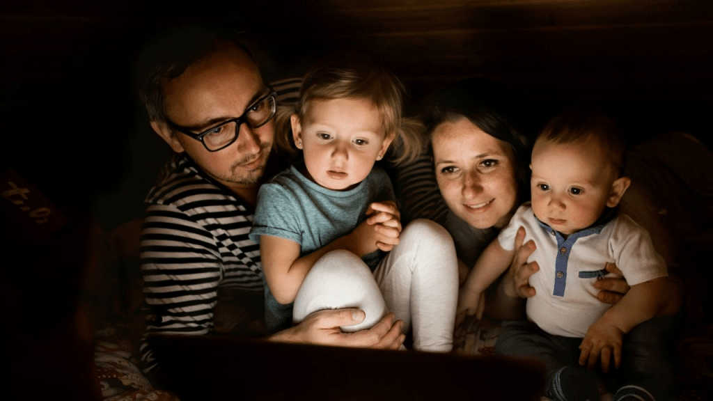 A man, a woman, and two small children are sitting close together in the dark, looking at a laptop with a glowing screen.
