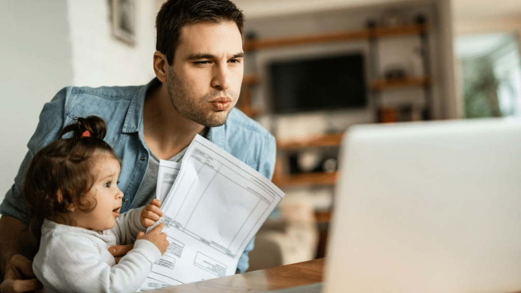 A man and a baby sit together, looking at a laptop with papers in their hands.