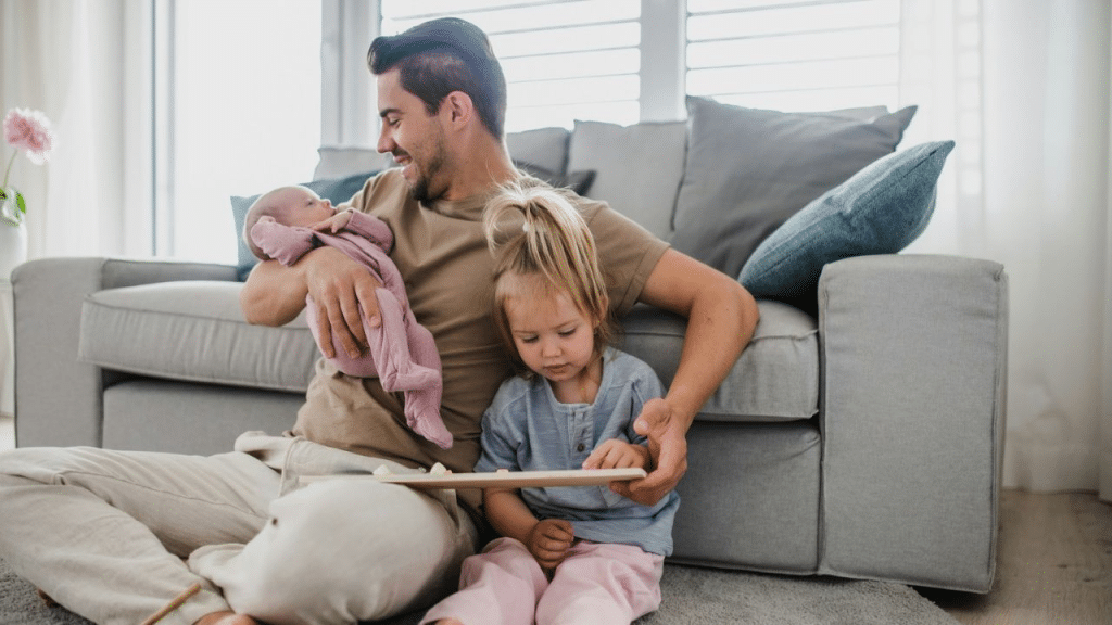 A man sits on the floor with a baby in his arms, and a young girl sits beside him, playing with a toy.